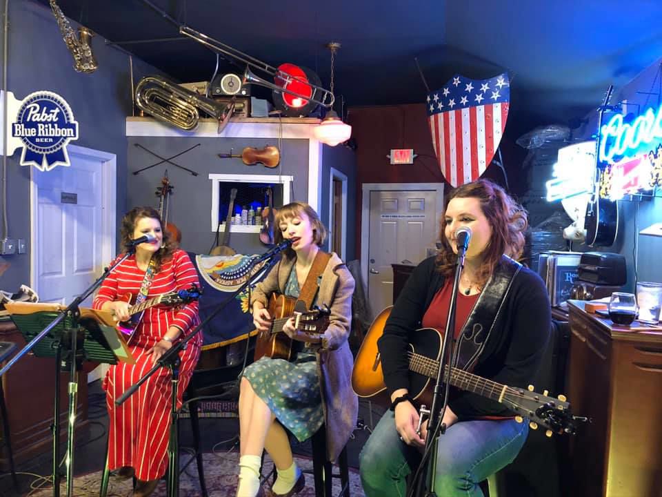 Three women sit while holding guitars as they sing into microphones